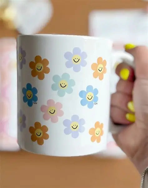 White mug with colorful flower designs featuring smiley faces, held by a person.
