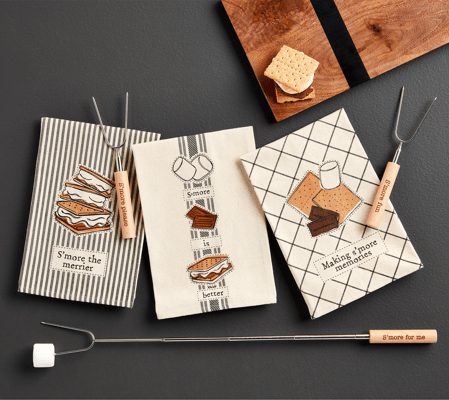 Set of s'mores-themed kitchen towels and a marshmallow roasting stick on a dark surface.