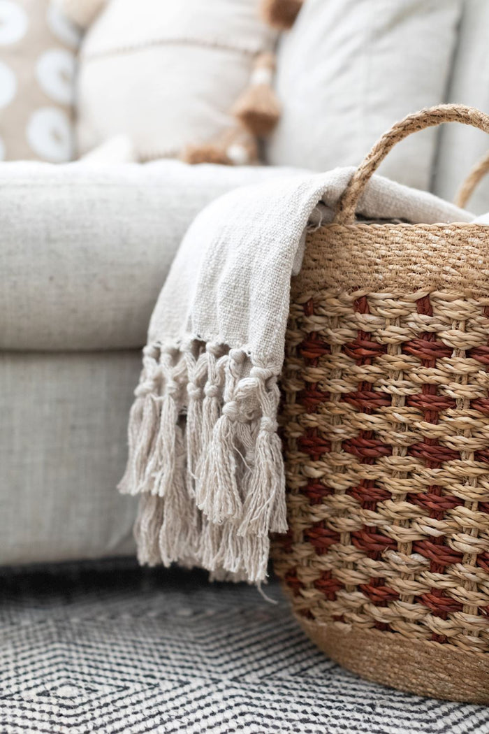 Beige throw blanket with tassels on a textured surface next to a woven basket.